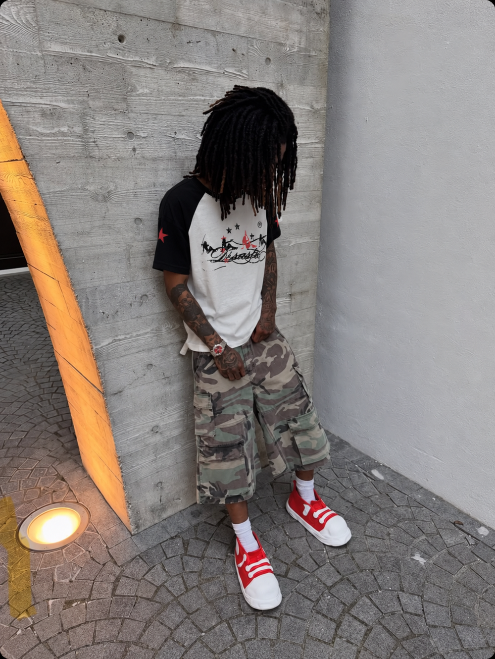 Désastre T-shirt porté avec un short camouflage contre un mur en béton.